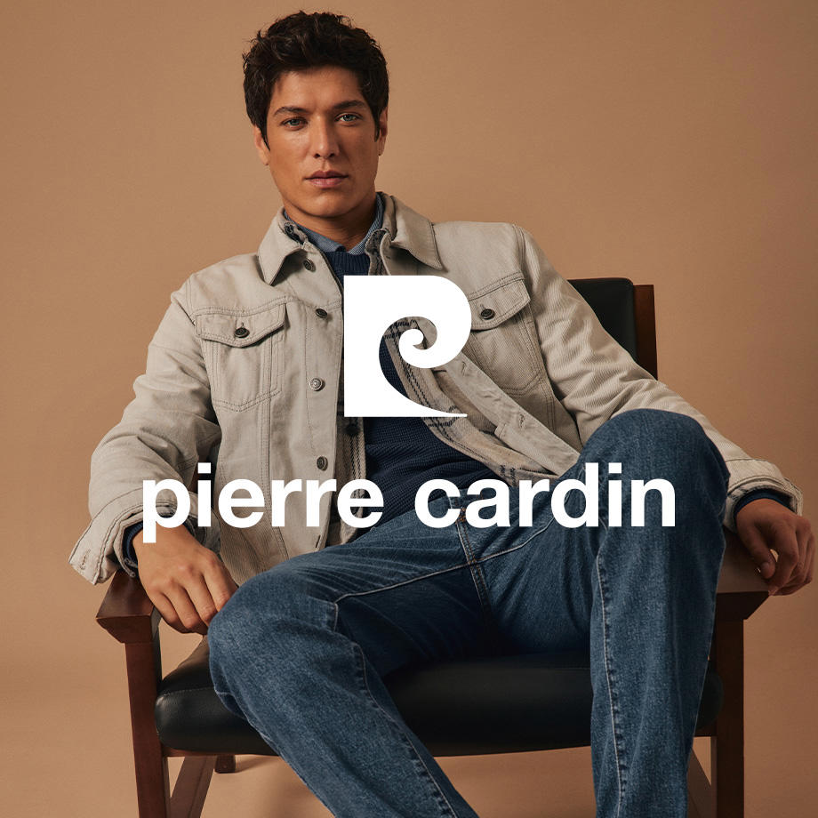 Ein Markenbild von der Marke "Pierre Cardin". Ein Mann sitzt lässig auf einem Holzstuhl in einem Innenraum mit braunem Hintergrund. Er trägt eine beige Jeansjacke, eine dunkle Jeans sowie einen dunkelblauen Pullover mit Hemd darunter – ein eleganter, moderner Look von Pierre Cardin in warmer Indoor-Atmosphäre.