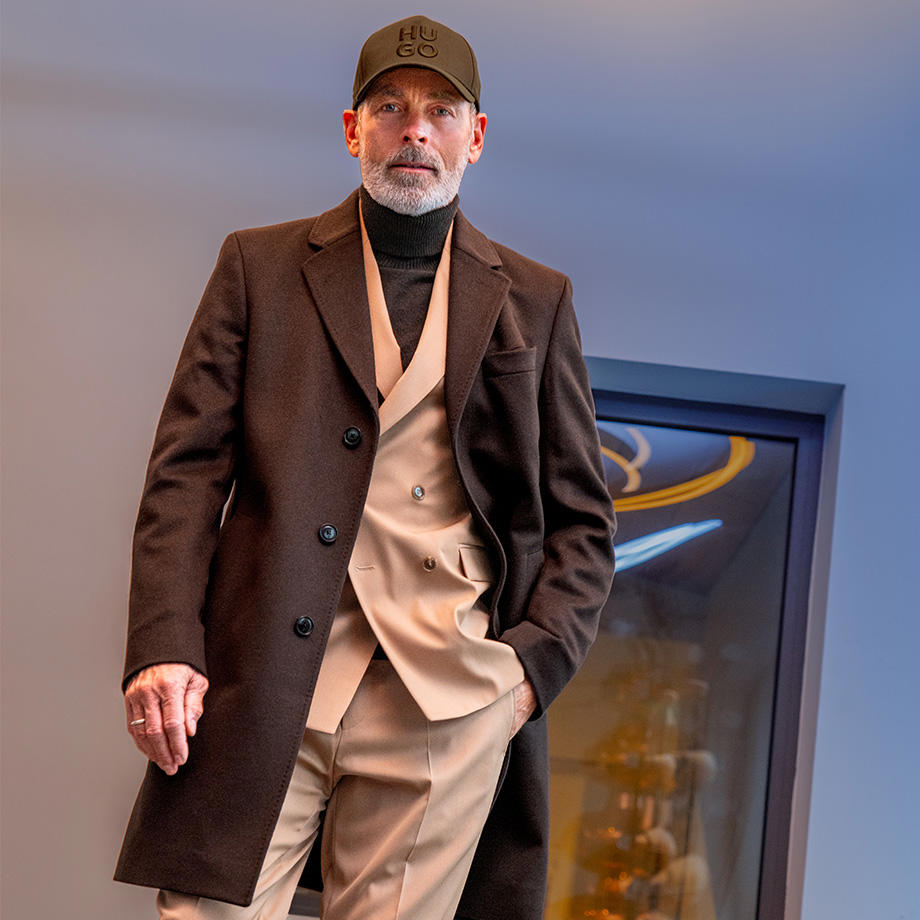Ein männliches Model zeigt einen modernen Layering-Look, der aus einem grauen Strickpullover, einem grauen Sakko mit Kapuze und einem beige-grauen Mantel besteht, waehrend das Model mit einer Hand laessig an einer Wand lehnt und in einer urbanen, aeusseren Umgebung posiert.