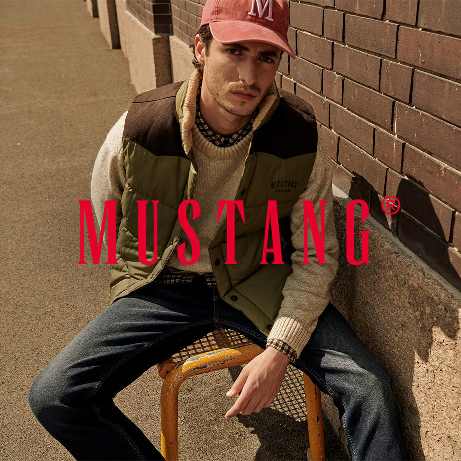 Ein Markenbild von der Marke "Mustang". Ein Mann sitzt lässig auf einem gelben Metallstuhl an einer Backsteinwand. Er trägt eine olivgrüne Steppweste mit „Mustang“-Logo, einen beigen Strickpullover, eine Jeans und eine rote Cap mit weißem „M“. Der Look wirkt entspannt und herbstlich-urban.