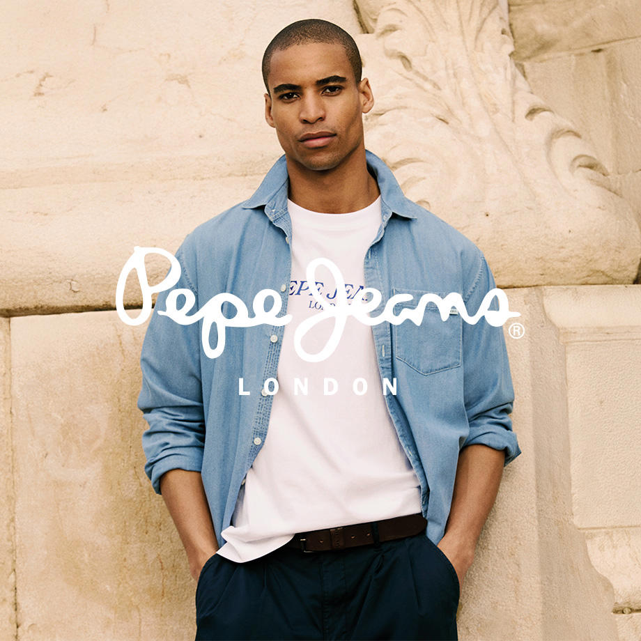 Ein Markenbild von der Marke "Pepe Jeans". Ein Mann lehnt entspannt an einer hellen Wand in der Sonne. Er trägt ein offenes, hellblaues Denim-Hemd über einem weißen T-Shirt mit Pepe Jeans Logo sowie eine dunkelblaue Stoffhose – ein lässiger, sommerlicher Look mit urbanem Flair.