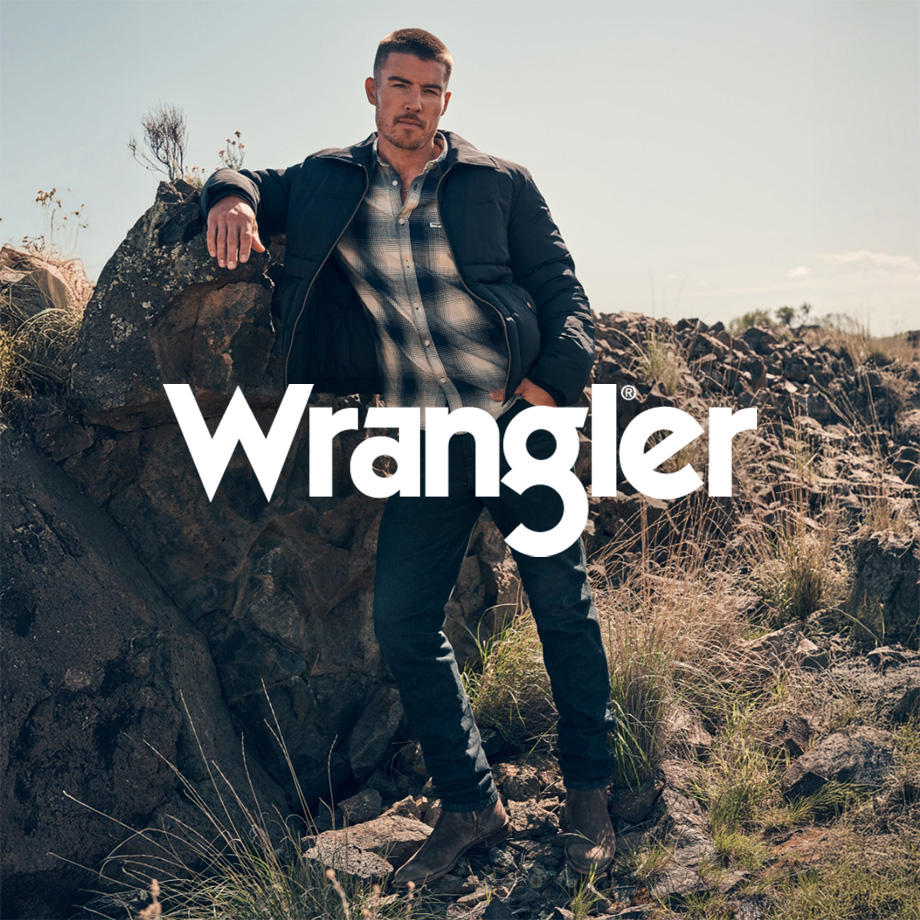 Ein Markenbild von der Marke "Wrangler". Ein Mann lehnt entspannt an einem großen Stein in sonniger, natürlicher Umgebung mit Wiese und verstreuten Felsen. Er trägt ein weiß-blau kariertes Hemd unter einer dunklen Übergangsjacke, dazu eine dunkle Jeans und halbhohe Stiefel – ein robuster, lässiger Wrangler-Look im Outdoor-Stil.