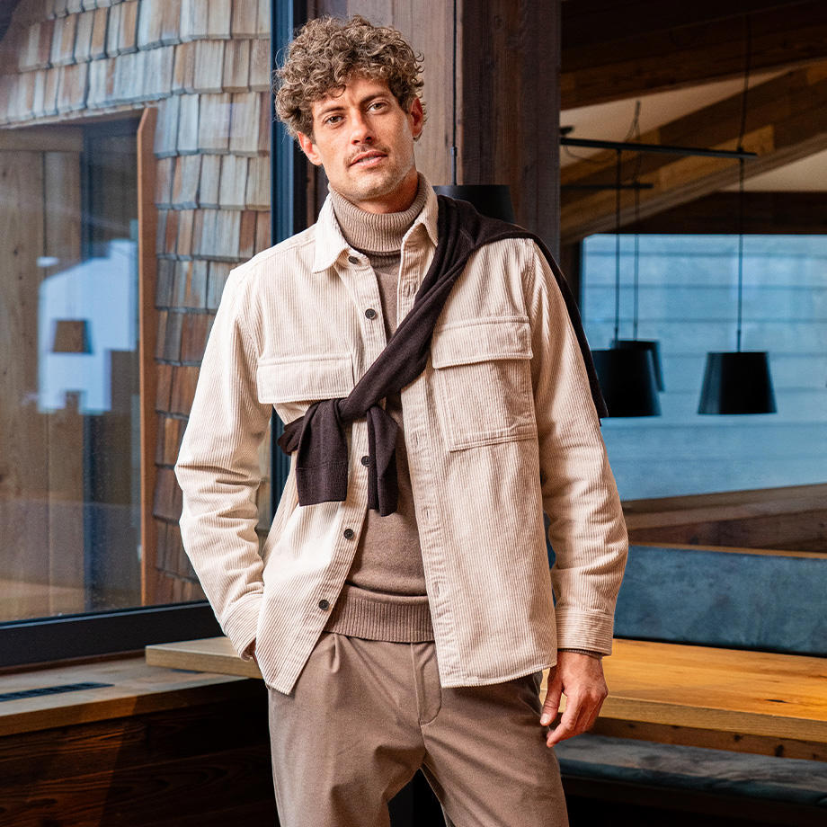 Ein männliches Model steht im Bahnpark und praesentiert ein stilvolles Layering-Outfit, das aus einem gemusterten Hemd, einem blau-beige gestreiften Pullover, einer blauen Weste und einem beigen Overshirt besteht, das mit einer braunen Hose kombiniert ist, wodurch der Look eine harmonische Farbpalette und laessige Eleganz ausstrahlt.