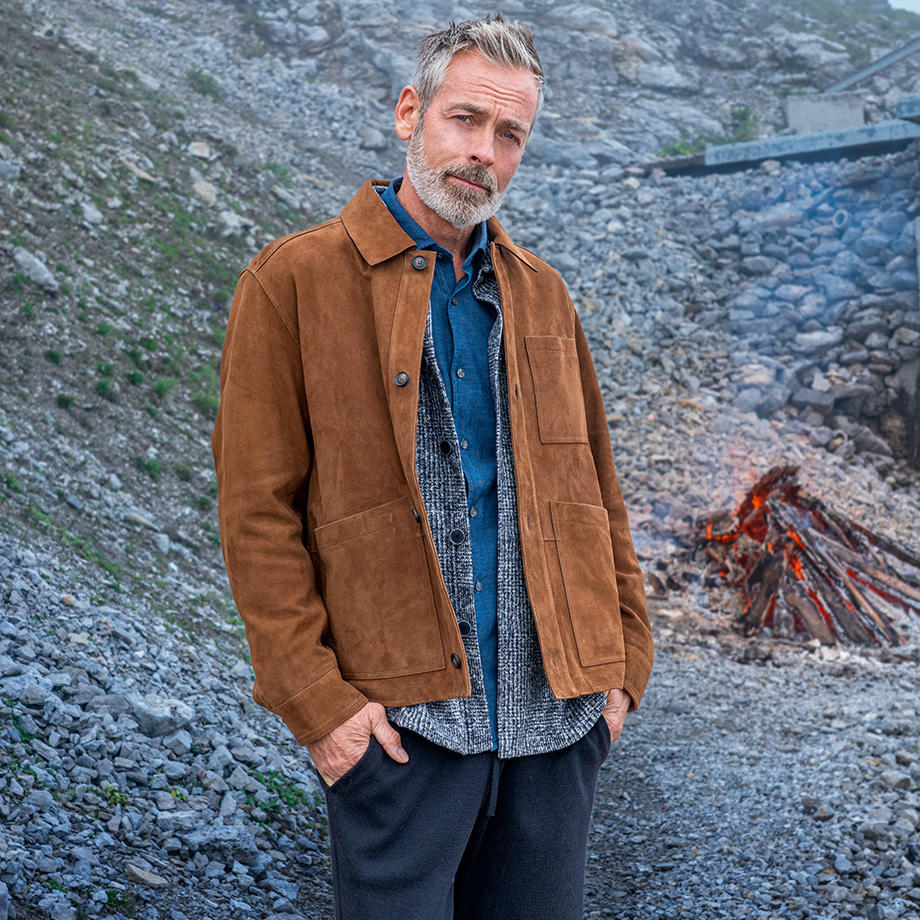Mann in brauner Wildlederjacke mit Denim-Hemd steht vor felsiger Berglandschaft.