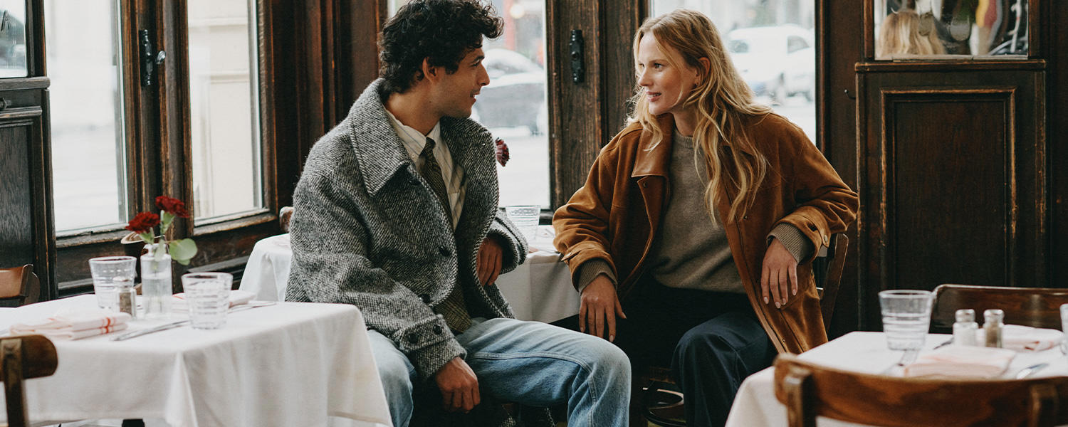 Ein Mann und eine Frau sitzen in einem stilvollen Café an einem gedeckten Tisch. Er trägt eine graue Gant Jacke über Hemd und Krawatte mit Jeans, sie kombiniert einen braunen Gant Mantel mit grauem Pullover. Urbaner, eleganter Look im entspannten Gespräch.