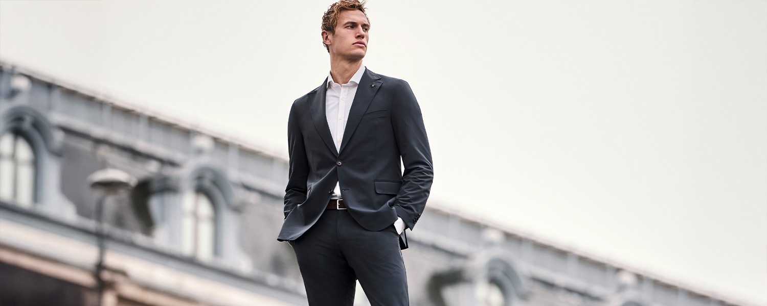 Mann trägt eleganten dunkelblauen Anzug mit weißem Hemd von Carl Gross und posiert selbstbewusst vor urbaner Architektur – stilvolle Business-Mode für Herren.