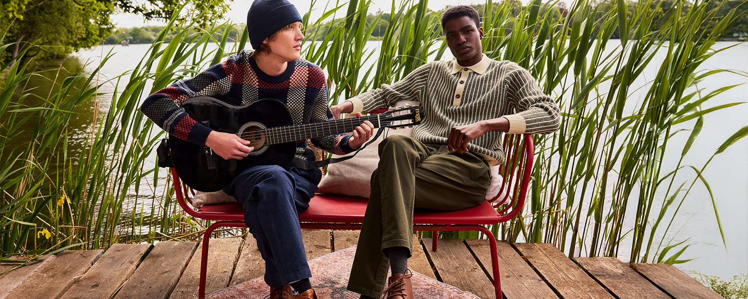 Zwei junge Männer sitzen auf einer roten Bank am Steg am See; einer spielt Gitarre im bunten Strickpullover und Mütze, der andere trägt einen beigen Strickpullover mit Knopfleiste – entspannte Outdoor-Mode im natürlichen Ambiente.