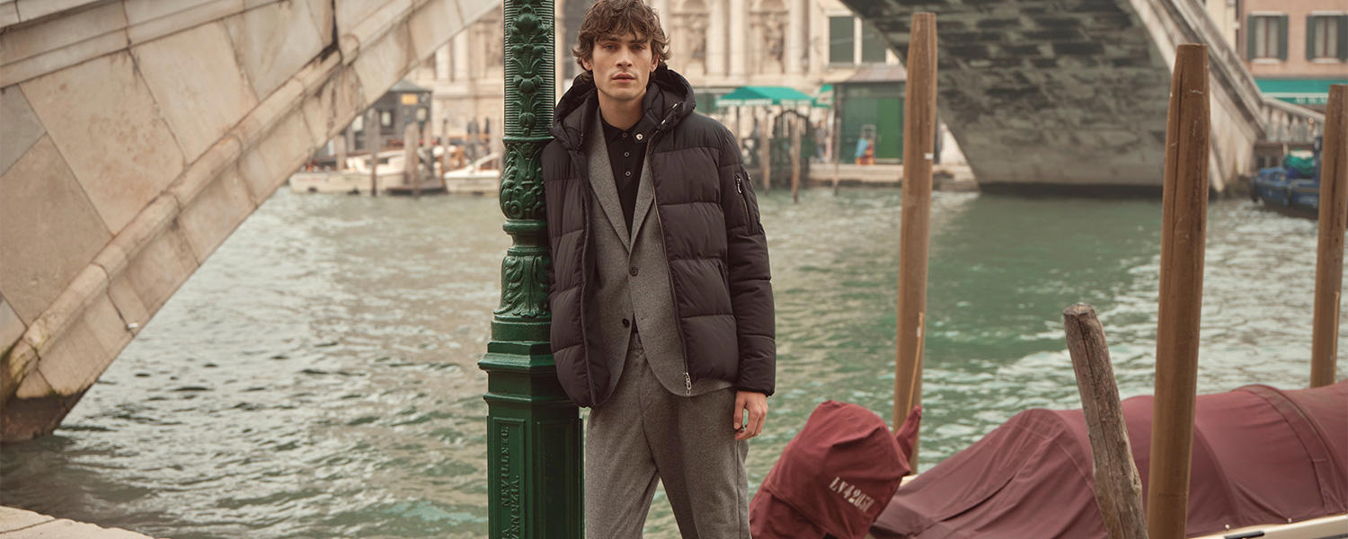 Mann trägt grauen Anzug mit schwarzer Steppjacke von bugatti und lehnt lässig an einer Laterne vor der Rialtobrücke in Venedig – elegante Herrenmode mit urbanem Flair.