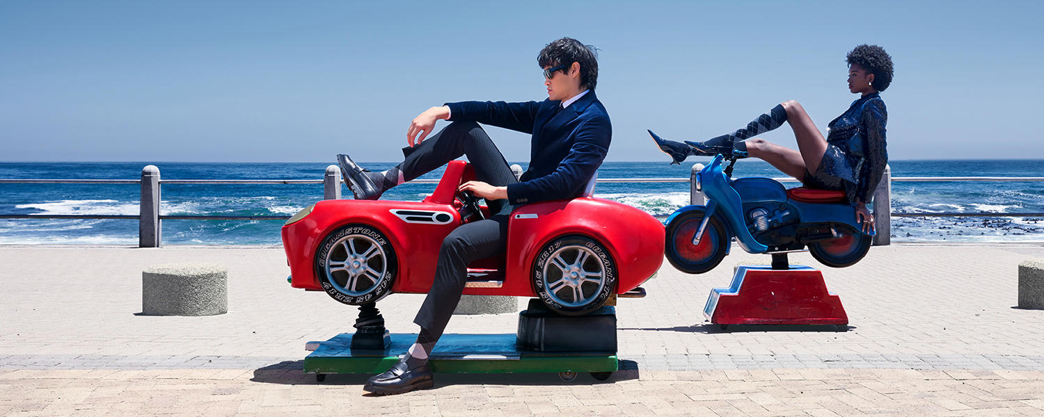 Mode-Shooting von Burlington am Meer: Ein Mann in elegantem Anzug sitzt lässig in einem roten Spielzeugauto, während eine Frau im glitzernden Mini-Outfit und kniehohen Strümpfen auf einem blauen Spielzeug-Motorrad posiert – stilvolle Burlington Mode mit Blick auf den Ozean.