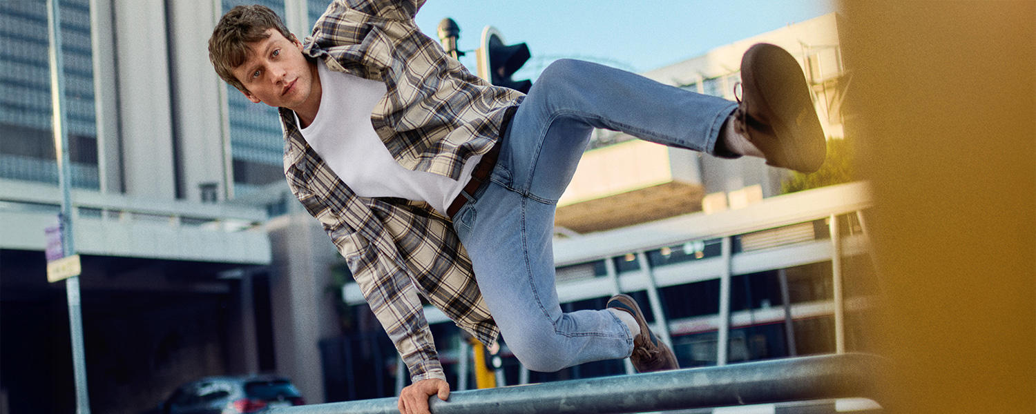 Junger Mann trägt kariertes Hemd, weißes T-Shirt und Jeans von camel active und springt sportlich über ein Geländer in urbaner Umgebung – lässige Herrenmode für Freizeit und Alltag.