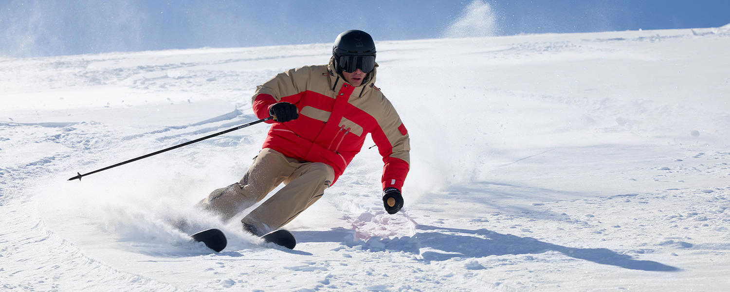 Skifahrer in beige-roter Skijacke und Hose von Bogner fährt dynamisch eine verschneite Piste hinab – exklusive Skibekleidung für höchste Performance.