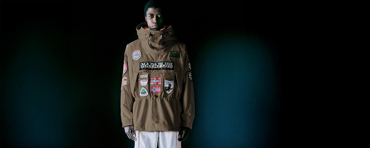 Mann trägt braune Napapijri Outdoor-Jacke mit auffälligen Patches und Logos vor dunklem Hintergrund.
