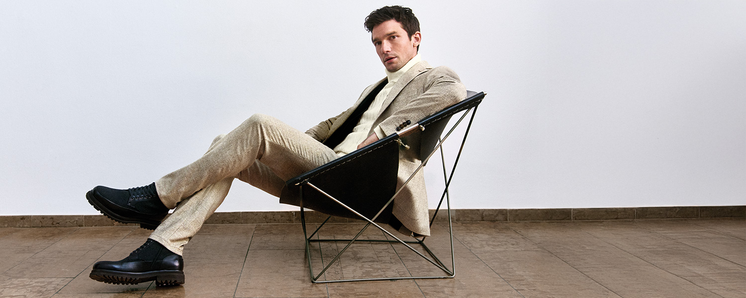 Mann sitzt entspannt in modernem Designerstuhl und trägt beigefarbenen Digel Anzug mit hellem Rollkragenpullover und schwarzen Schnürboots – stilvolle Businessmode für Herren.