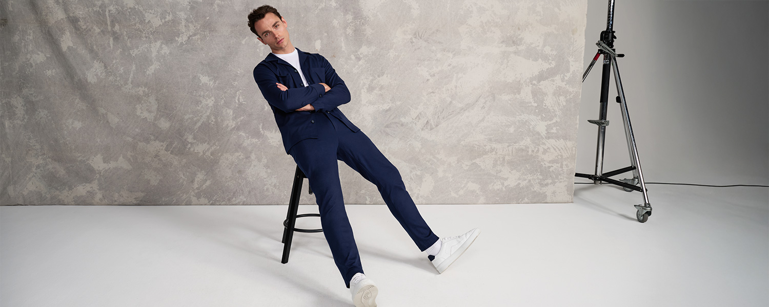 Mann sitzt lässig auf einem Hocker und trägt ein modernes, dunkelblaues Set aus Hemdjacke und Hose von Desoto mit weißen Sneakern – stilvolle Herrenmode im minimalistischen Studio-Setting.
