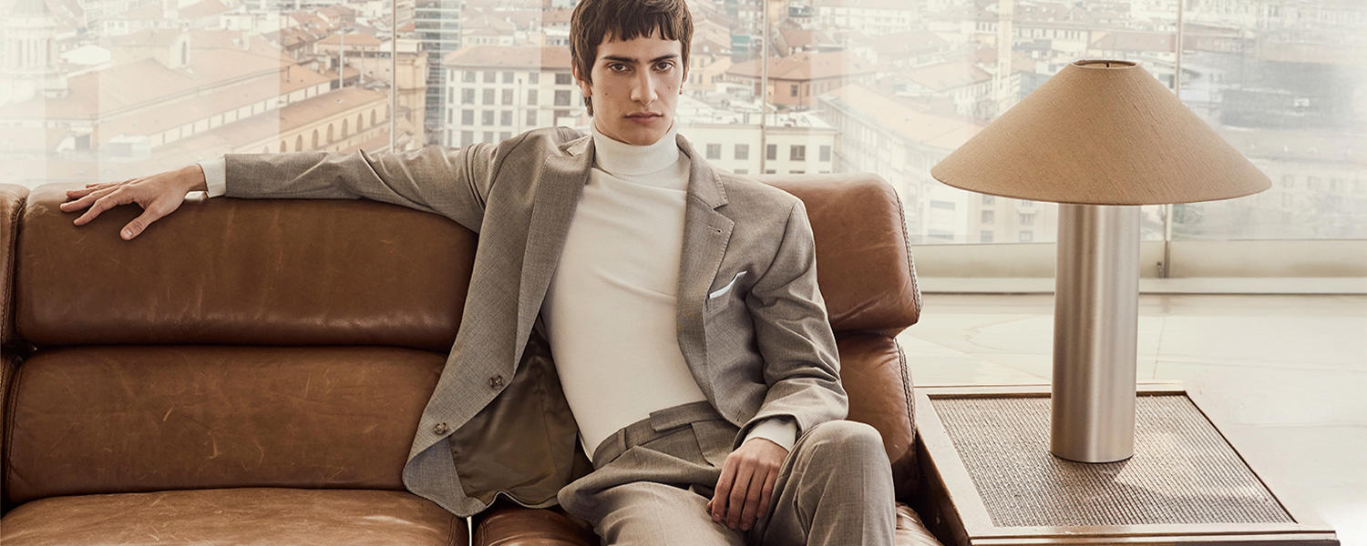 Mann sitzt entspannt auf einem Ledersofa in einem hellgrauen Cinque Anzug mit weißem Rollkragenpullover – elegante Herrenmode mit urbanem City-Flair.