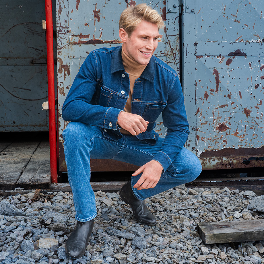 Ein Mann posiert im Freien vor einer rostigen Metalltür auf einem Schotterboden. Er trägt ein modisches Jeans-Outfit bestehend aus einer klassischen blauen Jeansjacke und einer passenden blauen Röhrenjeans. Unter der Jacke trägt er einen beigefarbenen Rollkragenpullover. Sein Look wird durch schwarze Leder-Chelsea-Boots abgerundet.