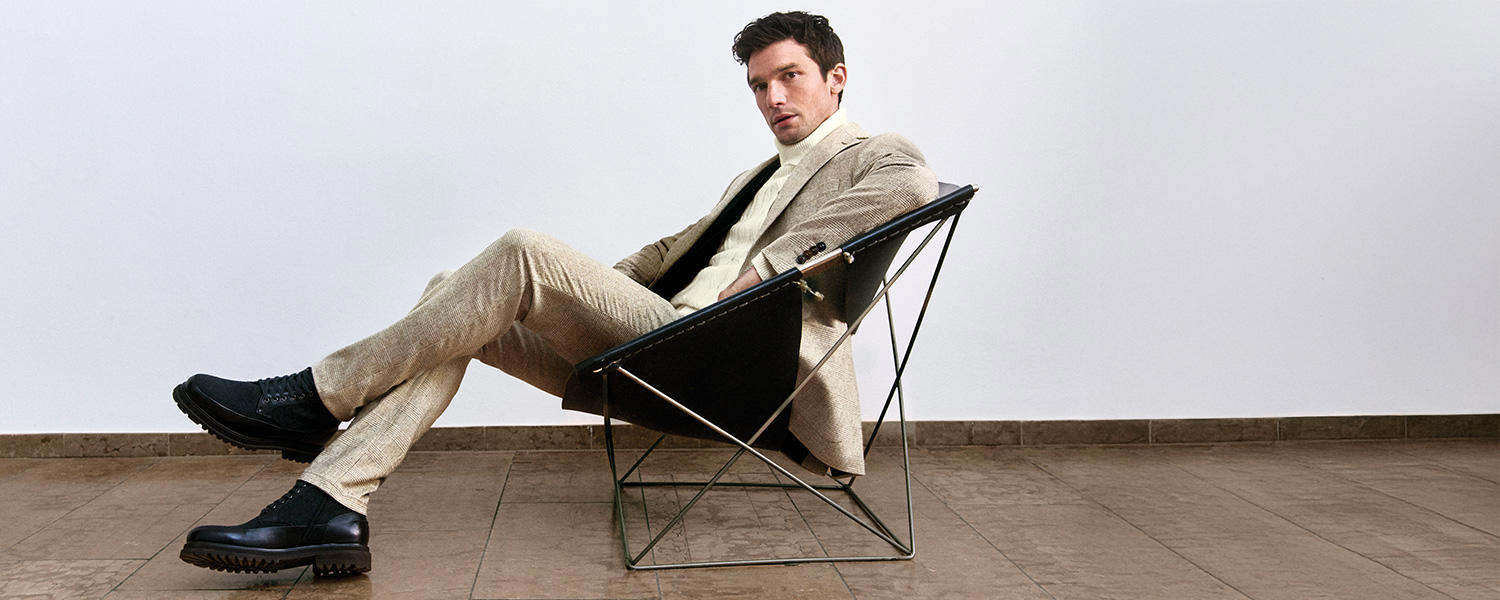 Mann sitzt entspannt in modernem Designerstuhl und trägt beigefarbenen Digel Anzug mit hellem Rollkragenpullover und schwarzen Schnürboots – stilvolle Businessmode für Herren.