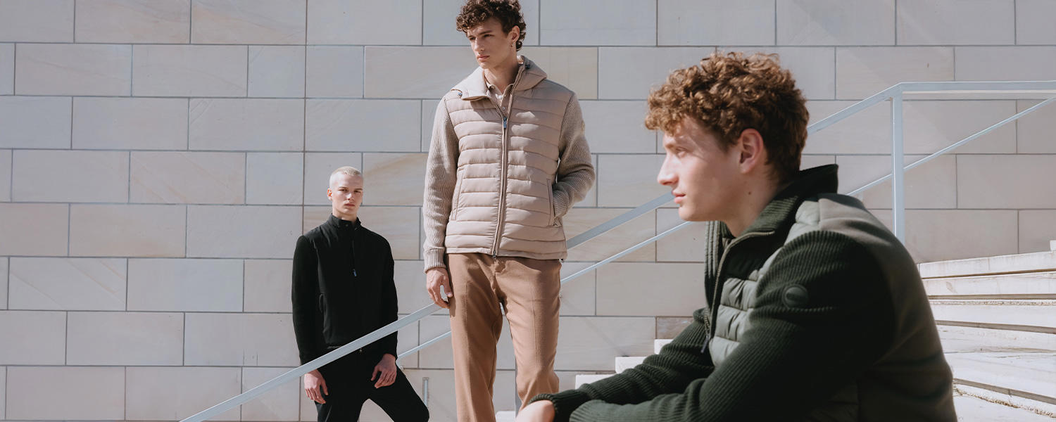 Drei Männer in modernen Steppwesten und sportlichen Jacken von Colmar posieren in urbanem Ambiente vor einer Steinwand – stylische Freizeitmode mit sportivem Charakter.