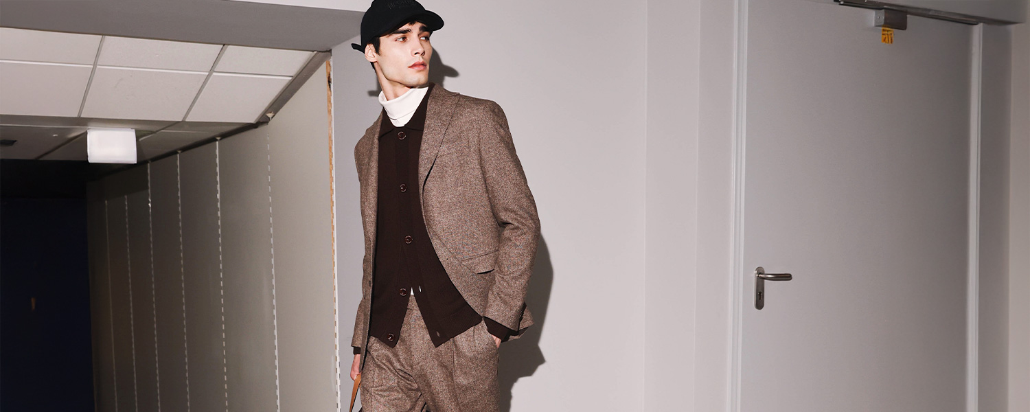 Mann trägt eleganten braunen Anzug mit dunkler Strickjacke und Rollkragen von Hechter Paris und kombiniert den Look mit schwarzer Cap – moderne Herrenmode mit urbanem Twist.