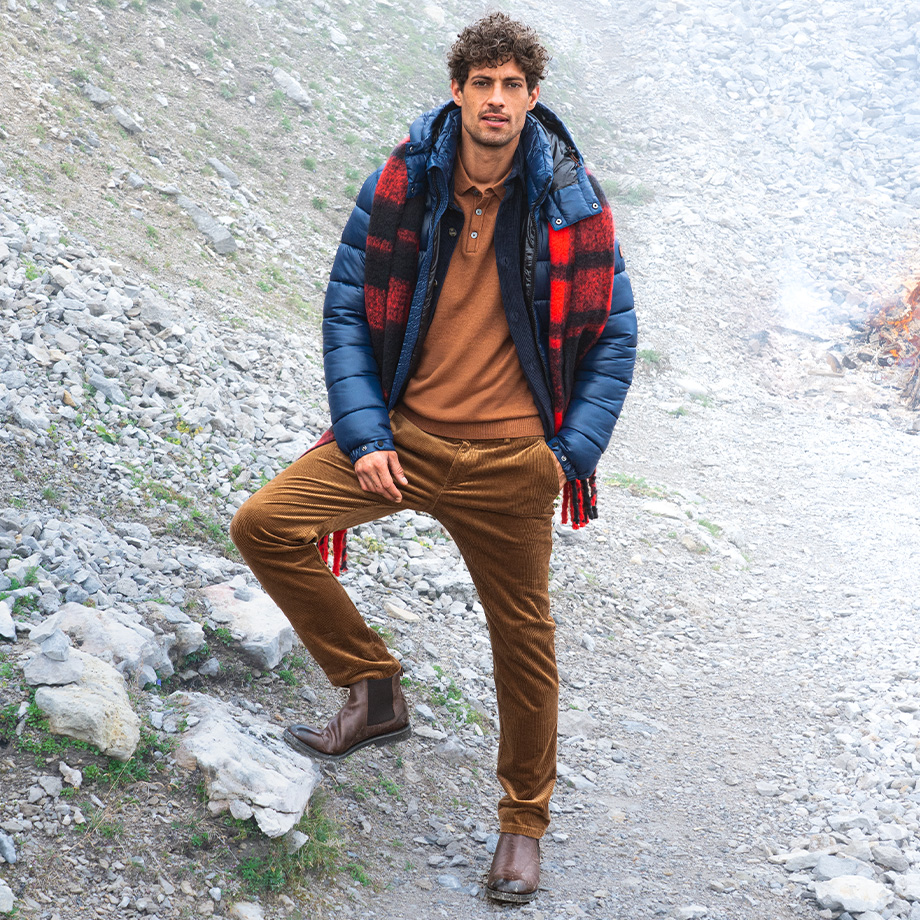 Mann in brauner Cordhose, blauer Steppjacke und Pullover in Caramel posiert auf steinigem Bergpfad – herbstlicher Outdoor-Look für modebewusste Männer.