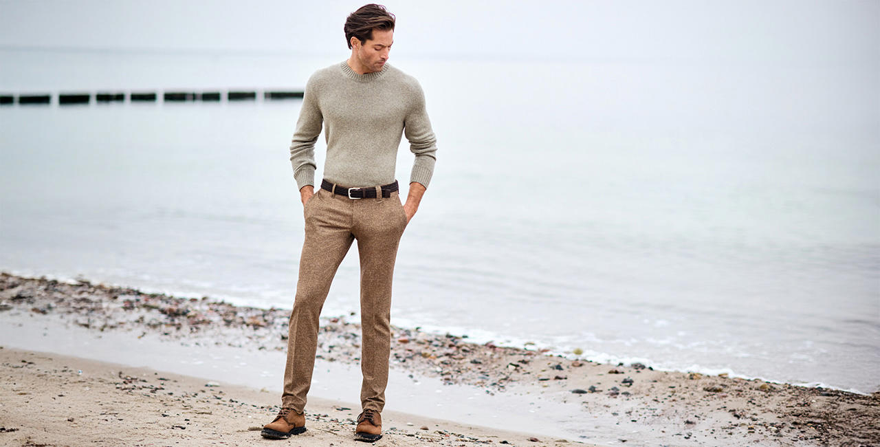 Elegant gekleideter Mann in beigefarbenem Strickpullover und brauner Stoffhose spaziert entspannt am Strand entlang – stilvolle Herrenmode für kühle Tage.