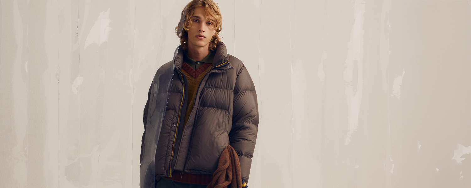 Junger Mann trägt eine voluminöse braune Daunenjacke von K-Way über mehrlagigem Strick-Look und hält einen Mantel in der Hand – stylische Wintermode für Herren.