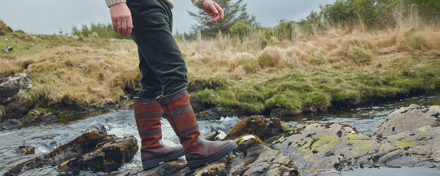 Mann trägt kniehohe braune Lederstiefel von Dubarry und steht in herbstlicher Naturkulisse auf Felsen am Fluss – hochwertige Outdoor-Schuhe für Herren.