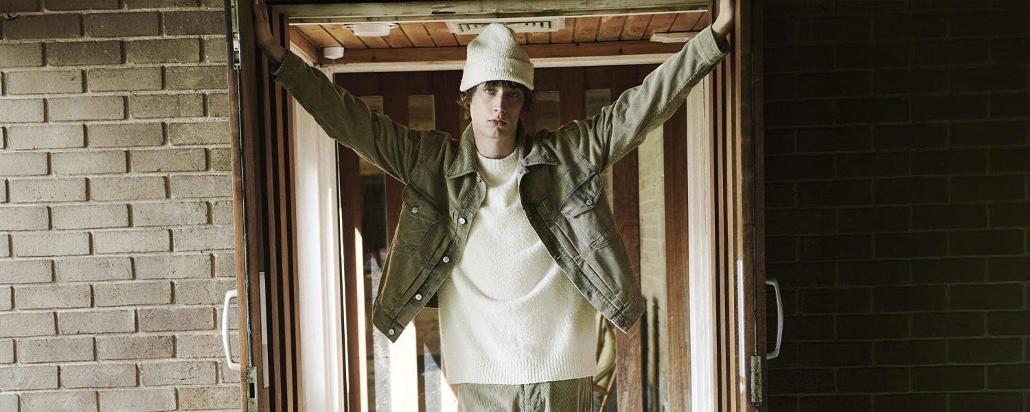 Junger Mann trägt Cordjacke mit passender Hose, weißen Strickpullover und Mütze von Denham und posiert lässig in einer Türöffnung – moderner Streetstyle im Retro-Look.