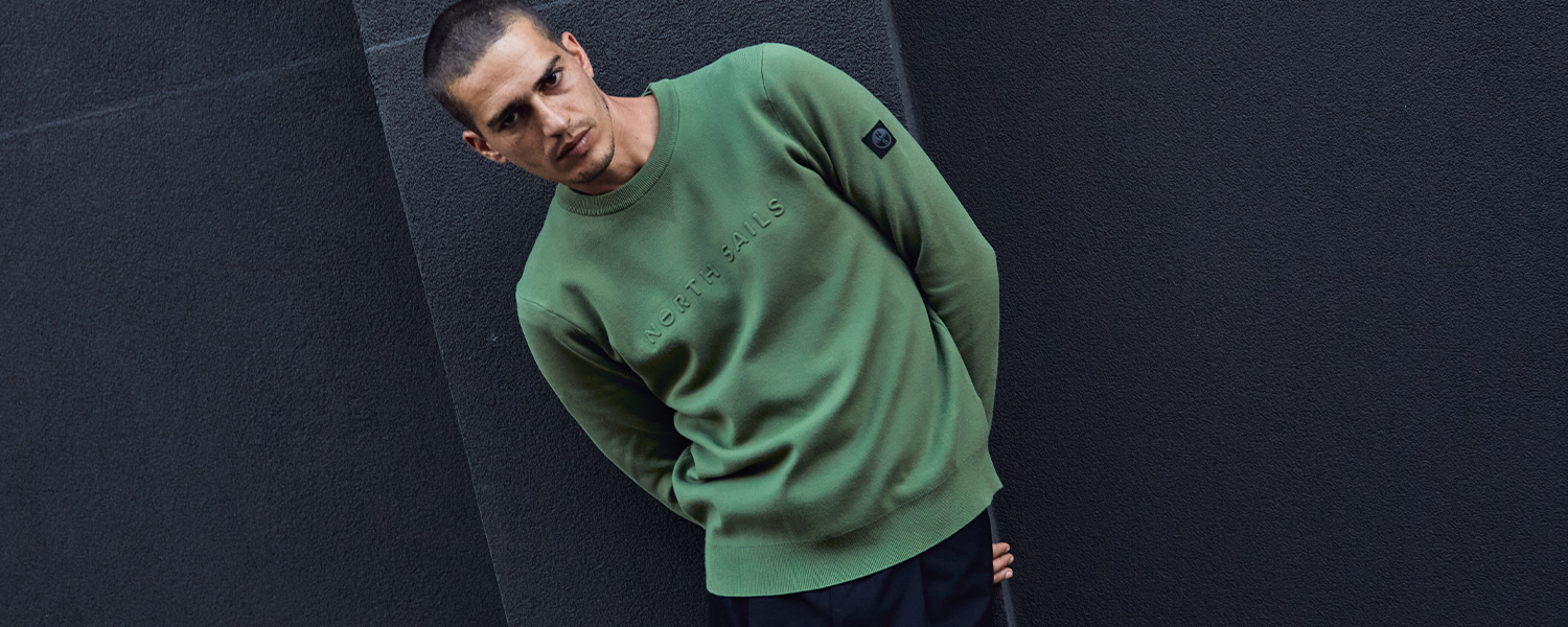 Mann trägt grünen North Sails Pullover mit Logo-Prägung vor dunkler Wand.