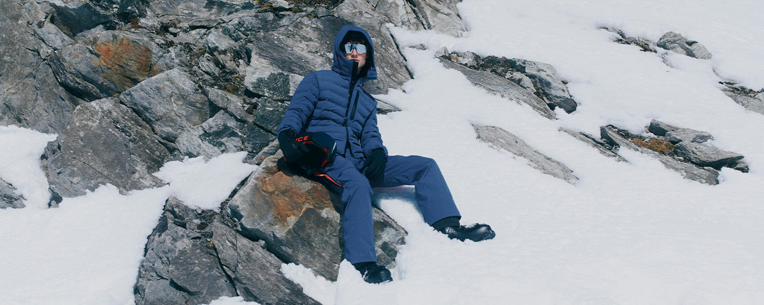 Mann in blauer Daunenjacke und Skihose von Bogner Fire+Ice sitzt entspannt auf Felsen im Schnee – hochwertige Wintermode für Outdoor und Ski.