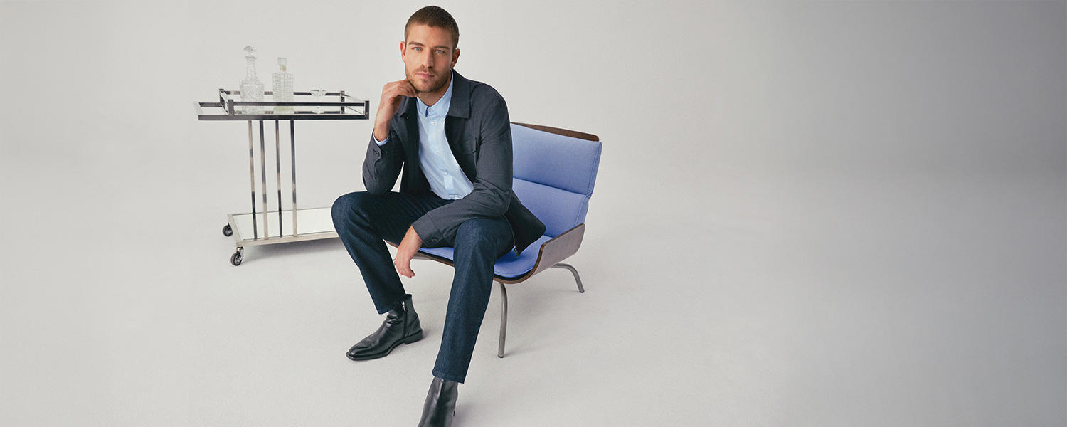 Ein Mann sitzt auf einem modernen blauen Sessel, trägt einen dunkelblauen Gardeur Anzug mit hellblauem Hemd und schwarzen Chelsea Boots, stilvoll präsentiert in einem minimalistischen Raum.
