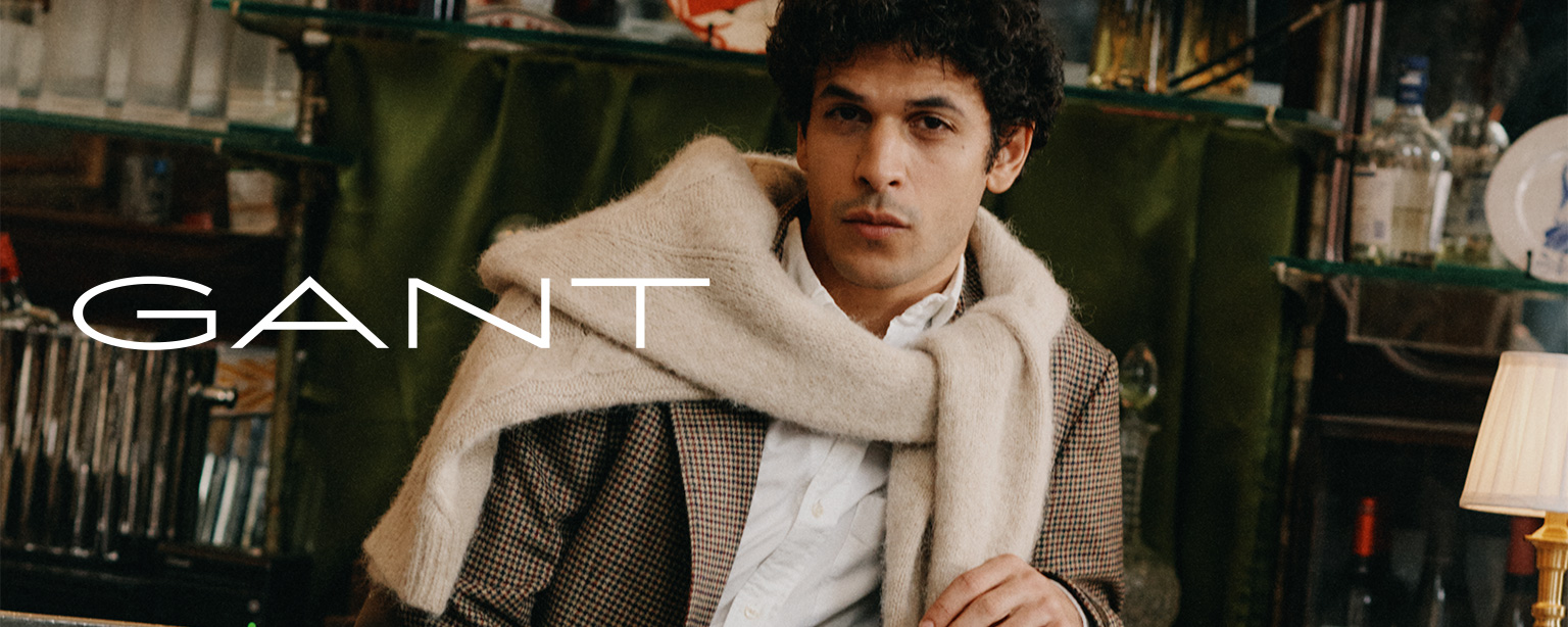 Lässig sitzender Mann in Karo-Blazer, weißem Hemd und über die Schultern gelegtem Pullover von GANT – zeitlose American Sportswear mit Stil.