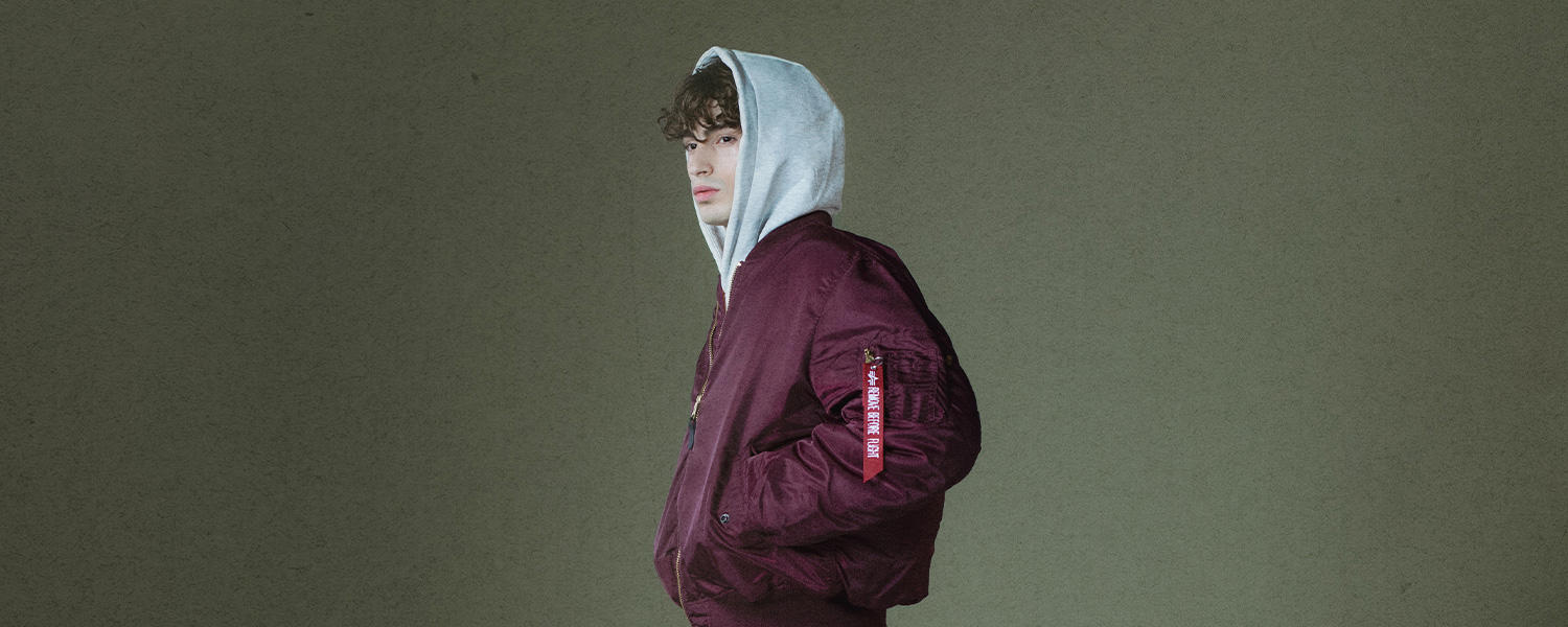 Das Bild zeigt einen jungen Mann mit dunkelrotem Blouson & grauem Hoodie vor grünem Hintergrund.