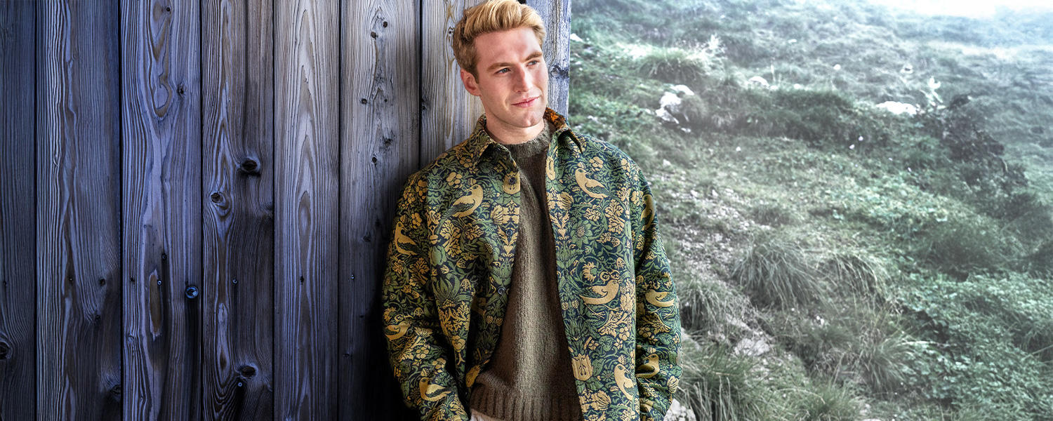 Auf dem Bild steht ein Mann draußen vor einer Holzwand und trägt einen olivgrünen Rundhalspullover unter einer gemusterten Jacke.