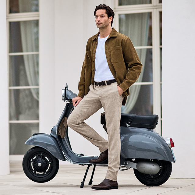 Mann in weißem T-Shirt mit brauner Overshirt-Jacke, beigefarbene Hose von Alberto und Chelsea-Boots, lässiger Casual-Look am Roller.