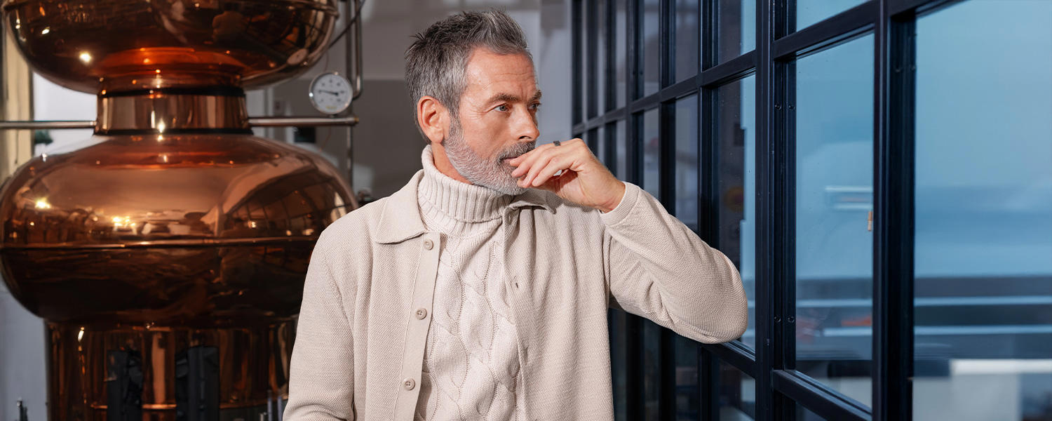 Stilvoll gekleideter Mann mit grauem Bart trägt einen beigen Cardigan über einem dicken Strickrollkragenpullover und blickt nachdenklich aus dem Fenster.