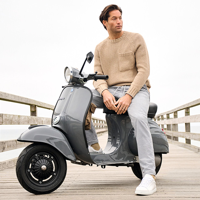 Mann in beigem Strickpullover mit Brusttasche und grauer Cordhose von Alberto, sitzend auf grauer Vespa am Steg