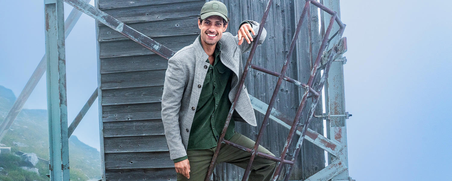 Grünes Cordhemd in extralanger Passform, getragen unter grauer Strickjacke mit Knopfleiste und Basecap im Outdoor-Setting.