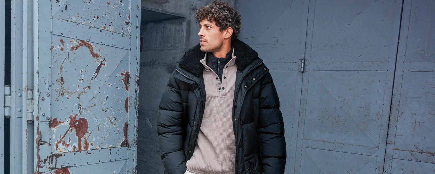 Auf dem Bild trägt ein Mann einen beigefarbenen Troyer mit halbem Zipper unter einer schwarzen Steppjacke.