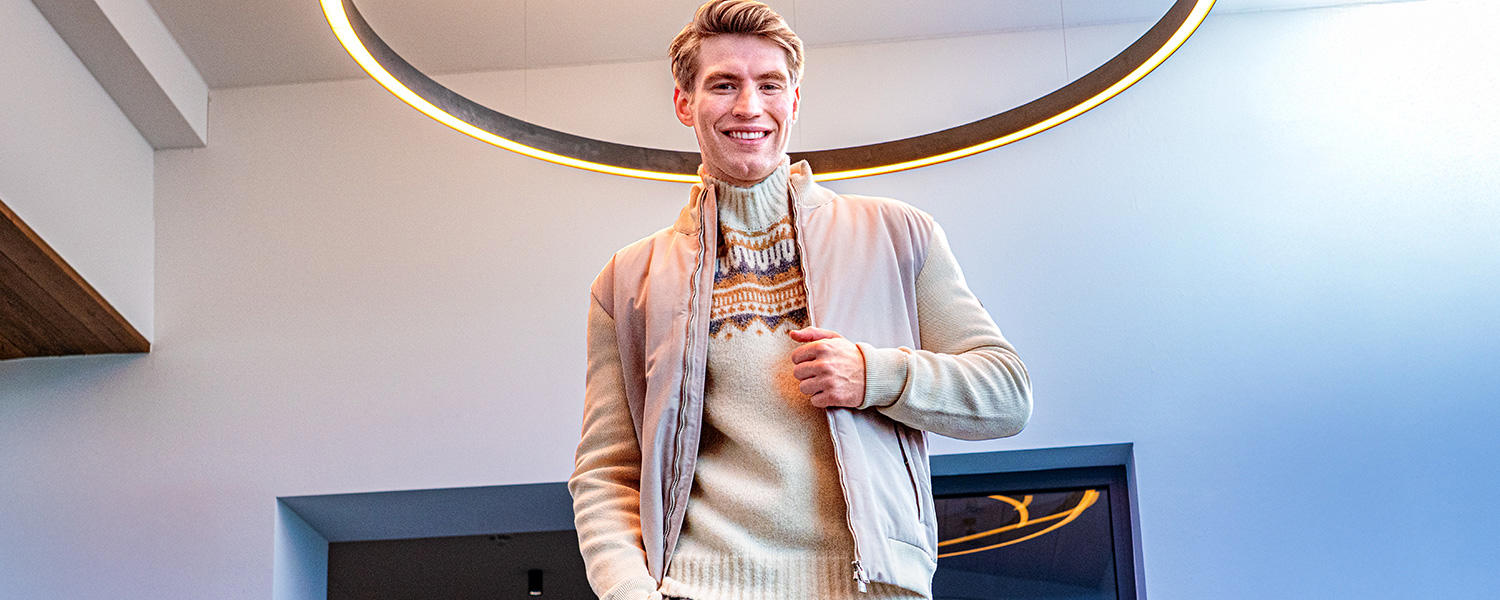Auf dem Bild trägt ein Mann einen hellen Rollkragenpullover mit Muster unter einer Jacke und lächelt in die Kamera.