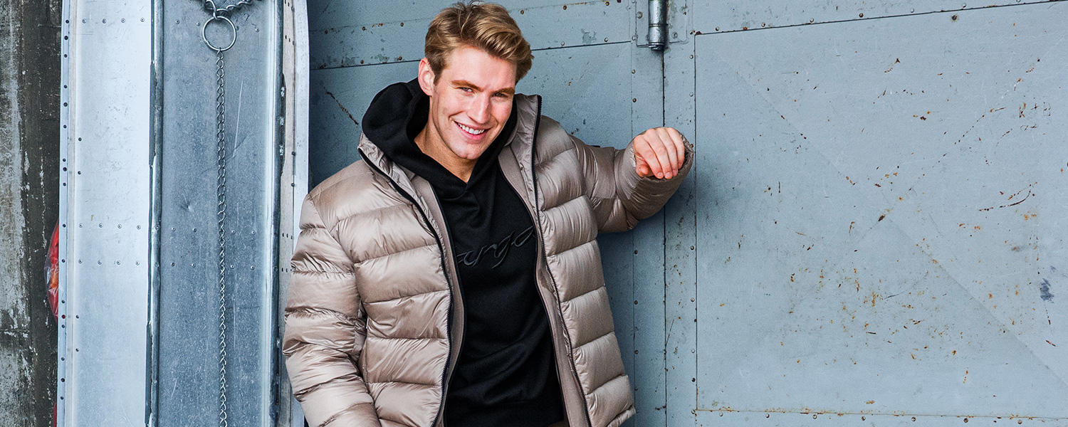 Auf dem Bild ist ein lächelnder Mann zu sehen, der einen beigefarbenen Daunenmantel über einem schwarzen Hoodie trägt.