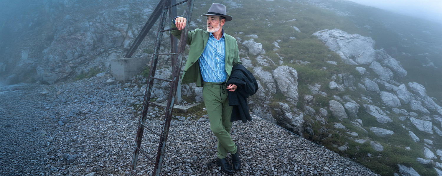 Mann im grünen Freizeitanzug mit blau gestreiftem Hemd und grauem Hut, leger getragen in alpiner Landschaft.