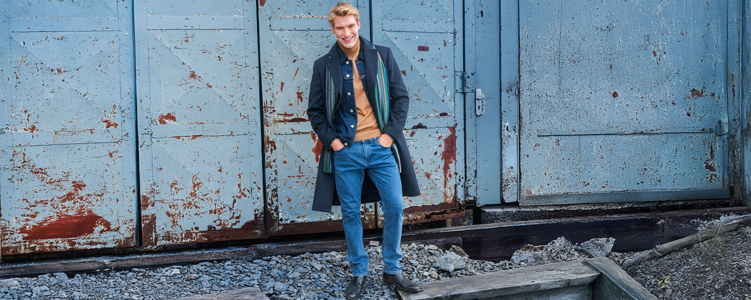 Mann in Jeans, Pullover und dunkelblauem Mantel steht vor einer alten Metalltür – lässiger Herbstlook für Herren.