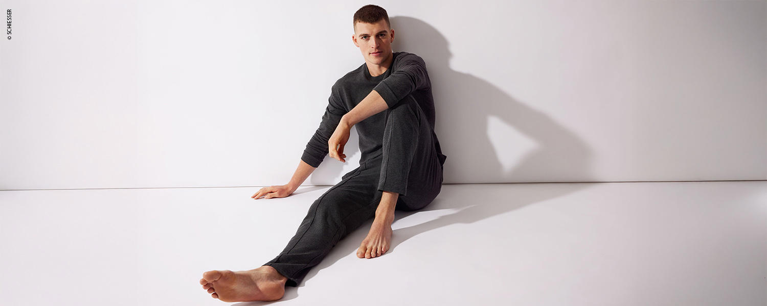 Ein Mann trägt eine dunkle Pyjama-Hose mit passendem Oberteil und sitzt entspannt auf dem Boden.