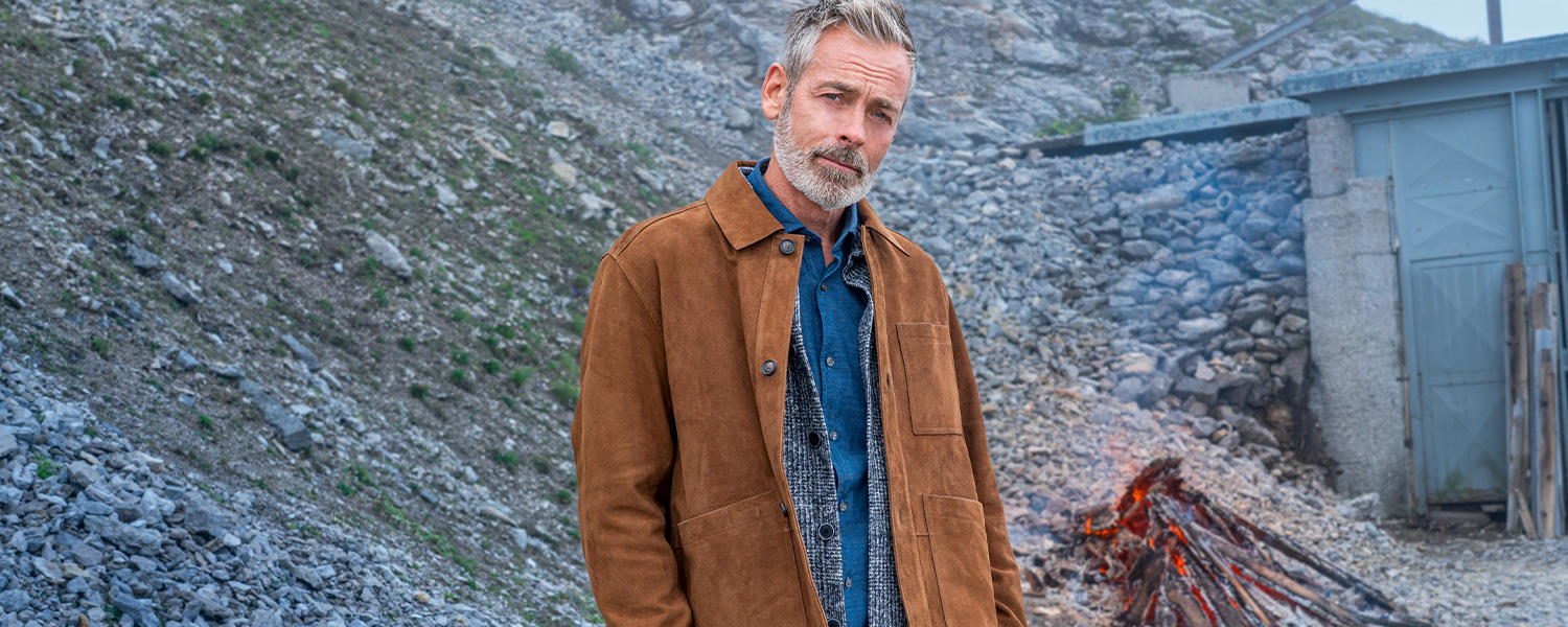 Blaues Jeanshemd in Kombination mit grauer Strickweste und brauner Wildlederjacke, getragen im Outdoor-Setting.