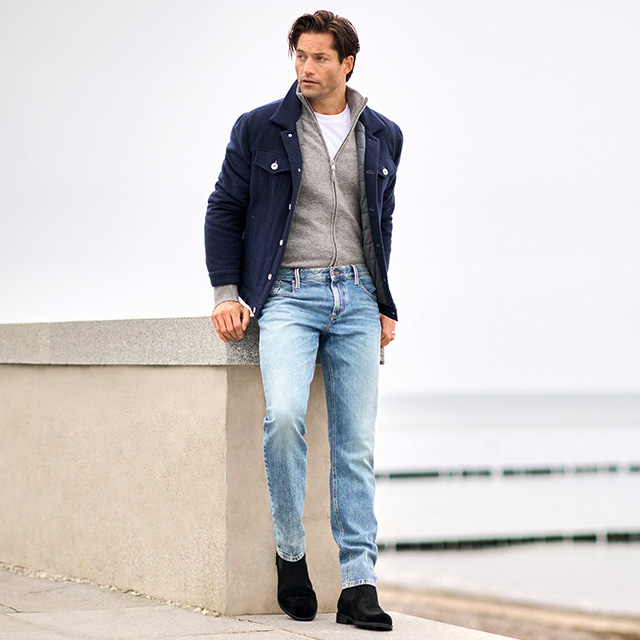 Mann in dunkelblauer Jacke mit grauem Zip-Cardigan und hellblauer Jeans von Alberto, lässig angelehnt am Geländer