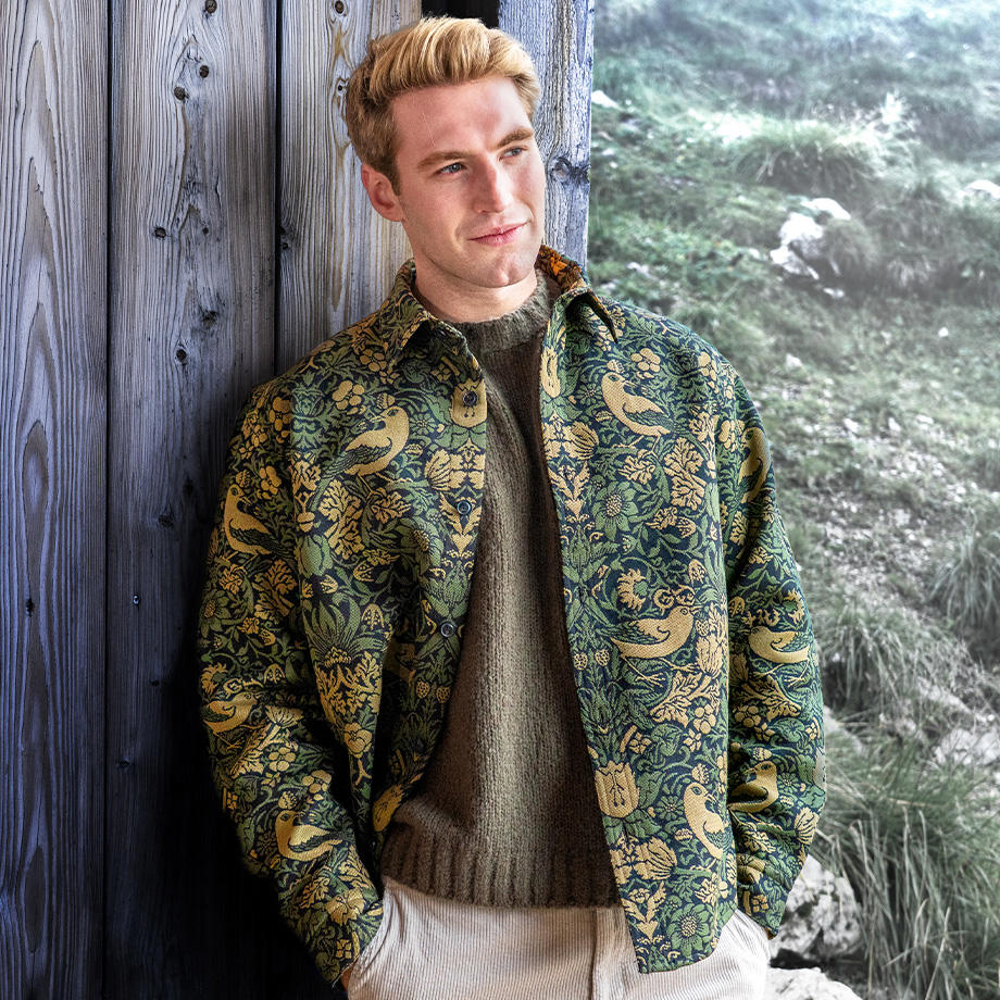 Ein Mann lehnt an einer Holzwand und trägt einen dicken grünen Strickpullover unter einem gelb-grün gemusterten Overshirt sowie eine beige Cordhose. Im Hintergrund sind Wiese und Felsen zu sehen. Das Outfit passt zur kühleren Witterung und vermittelt einen rustikalen, naturverbundenen Look.