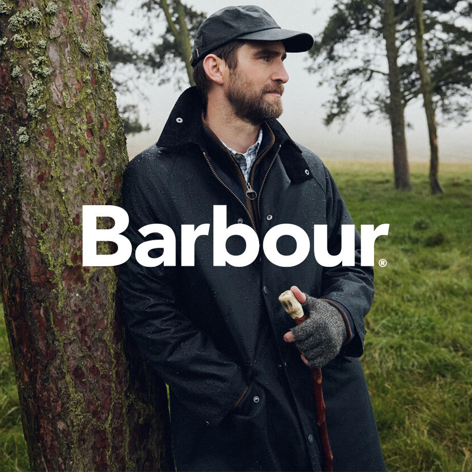 Ein Mann lehnt an einem Baum in einer ländlichen Umgebung und trägt eine klassische gewachste Jacke, Mütze und Handschuhe – perfekt für nasses Wetter. Die Szene vermittelt britisches Outdoor-Flair und ist Teil einer Kampagne der Traditionsmarke Barbour, deren Logo prominent im Vordergrund zu sehen ist.