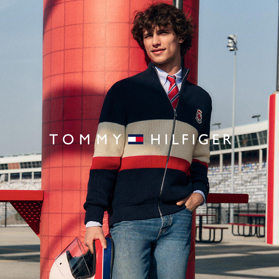 Mann in navyblauer Strickjacke mit rot-beigen Streifen, dazu Hemd und rote Krawatte von Tommy Hilfiger, lässig an eine Säule angelehnt mit Jeans. 