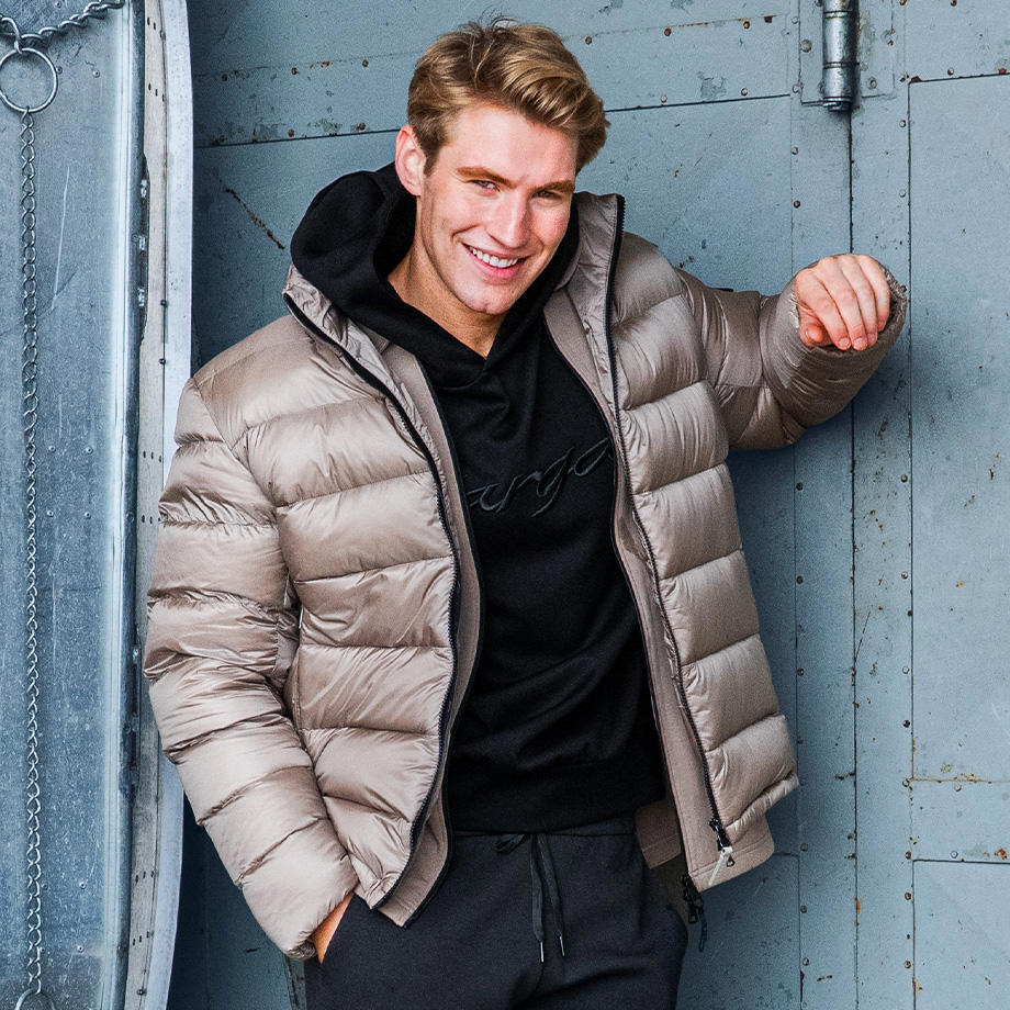 Ein Mann lehnt lässig an einer grauen Wand und trägt eine hellbeige Daunenjacke über einem schwarzen Hoodie. Das Outfit kombiniert urbanen Streetstyle mit wintertauglicher Funktionalität und vermittelt einen modernen, entspannten Look.