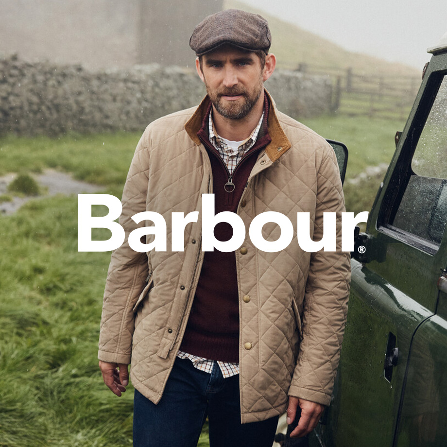 Mann in beiger Steppjacke mit braunem Pullover, kariertem Hemd und Schiebermütze von Barbour, stehend neben Geländewagen im Grünen